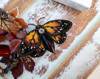 May include: A beaded butterfly brooch with orange, brown, and black wings. The brooch is on a wooden surface with a brown and white painted finish.