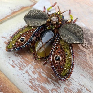 May include: A beaded and embroidered brooch in the shape of a beetle with a green, brown, and gold color scheme. The beetle has a large, clear gemstone in its center.