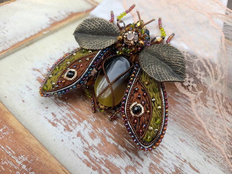 May include: A beaded and embroidered brooch in the shape of a beetle with a green, brown, and gold color scheme. The beetle has a large, clear gemstone in its center.