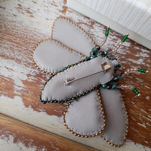 May include: A handmade gray leather butterfly brooch with gold beaded edges and iridescent green and clear beaded antennae. The brooch has a pin back for easy attachment to clothing or accessories. The butterfly's wings are a soft gray color.
