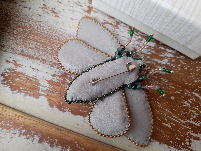 May include: A handmade gray leather butterfly brooch with gold beaded edges and iridescent green and clear beaded antennae. The brooch has a pin back for easy attachment to clothing or accessories. The butterfly's wings are a soft gray color.