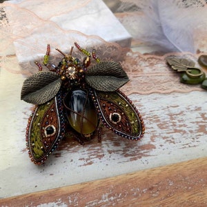 May include: A beaded insect brooch with green, brown, and gold accents. The insect has a large, clear, yellow-brown gemstone in its center. The brooch is on a white wooden surface.