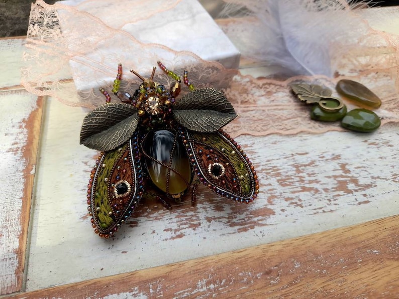 May include: A beaded insect brooch with green, brown, and gold accents. The insect has a large, clear, yellow-brown gemstone in its center. The brooch is on a white wooden surface.