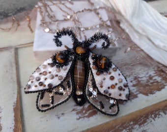 May include: A handcrafted moth brooch featuring a central brown striped stone body, two tiger eye stone accents, and white fabric wings embellished with small brown and grey beads. The edges are outlined with black seed beads and black fringe.