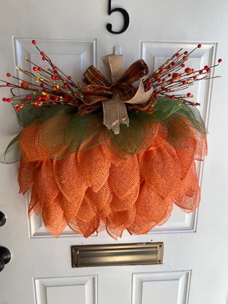 Op de afbeelding: Een herfst-deurkrans met oranje gaas, groene bladeren en rode bessen. In het midden zit een bruine jute strik met een geruit lint.