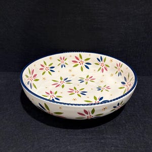May include: A round ceramic bowl with a cream-colored base featuring a repeating pattern of multi-colored floral motifs in shades of pink, blue, and green. The rim of the bowl is finished with a dark blue textured edge and small dots.