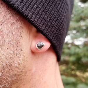 May include: A close-up of a person's ear wearing a small, silver earring with a black, faceted gemstone.