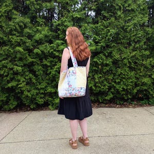 May include: A floral patterned tote bag with yellow panels is carried over the shoulder. The bag has a white strap. A person wearing a black dress carries the bag. The background is a dense green hedge.