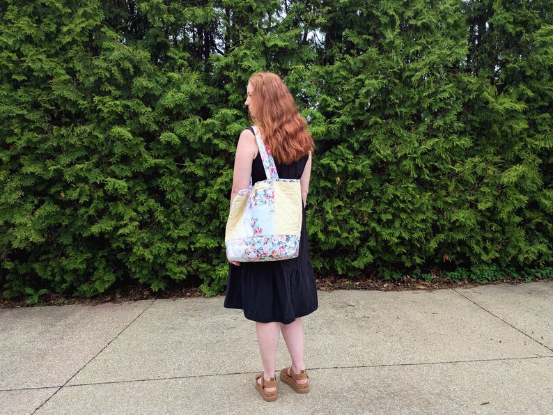 May include: A floral patterned tote bag with yellow panels is carried over the shoulder. The bag has a white strap. A person wearing a black dress carries the bag. The background is a dense green hedge.