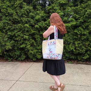 May include: A quilted tote bag with floral and light blue panels. The bag has a yellow quilted centre and floral patterned straps. The person is wearing a black dress and brown sandals.