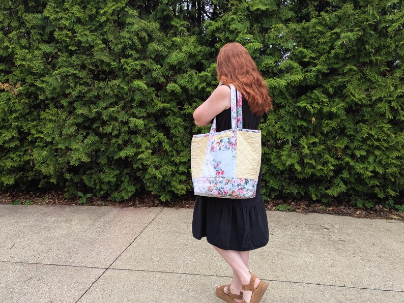 May include: A quilted tote bag with floral and light blue panels. The bag has a yellow quilted centre and floral patterned straps. The person is wearing a black dress and brown sandals.