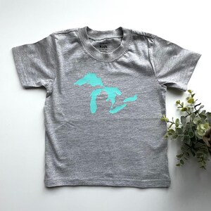 May include: A grey short-sleeved t-shirt with a turquoise outline of the Great Lakes on the front. The shirt has a tag that says "GarbKids" and "Kids".