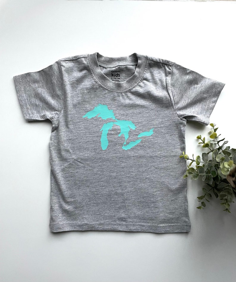 May include: A grey short-sleeved t-shirt with a turquoise outline of the Great Lakes on the front. The shirt has a tag that says "GarbKids" and "Kids".