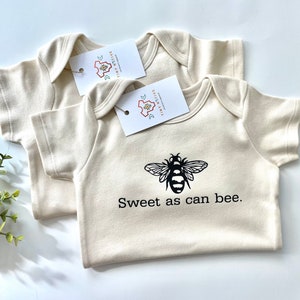 May include: Two cream-colored baby onesies with short sleeves. One onesie has a black bee graphic and the text "Sweet as can bee." The other onesie is plain.
