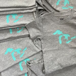 May include: A stack of grey t-shirts with a turquoise outline of the Great Lakes on each shirt. The top shirt has a white tag that says "Gerber Kids".