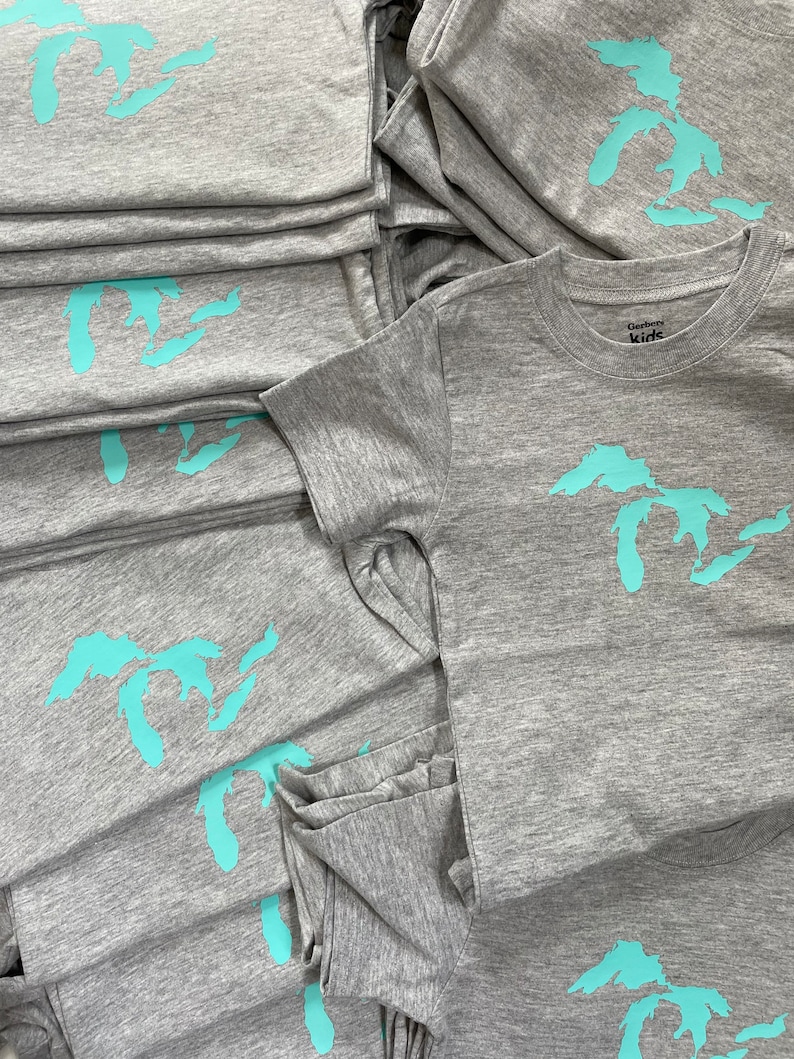 May include: A stack of grey t-shirts with a turquoise outline of the Great Lakes on each shirt. The top shirt has a white tag that says "Gerber Kids".