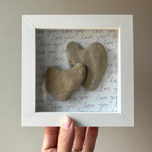 May include: A framed artwork featuring two heart-shaped stones. The stones are light brown and set against a background with handwritten "love you" text. The frame is white, creating a minimalist aesthetic.