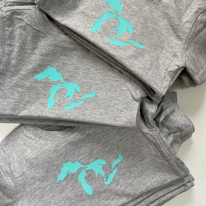 May include: A stack of grey t-shirts with a turquoise blue outline of the Great Lakes on each shirt.