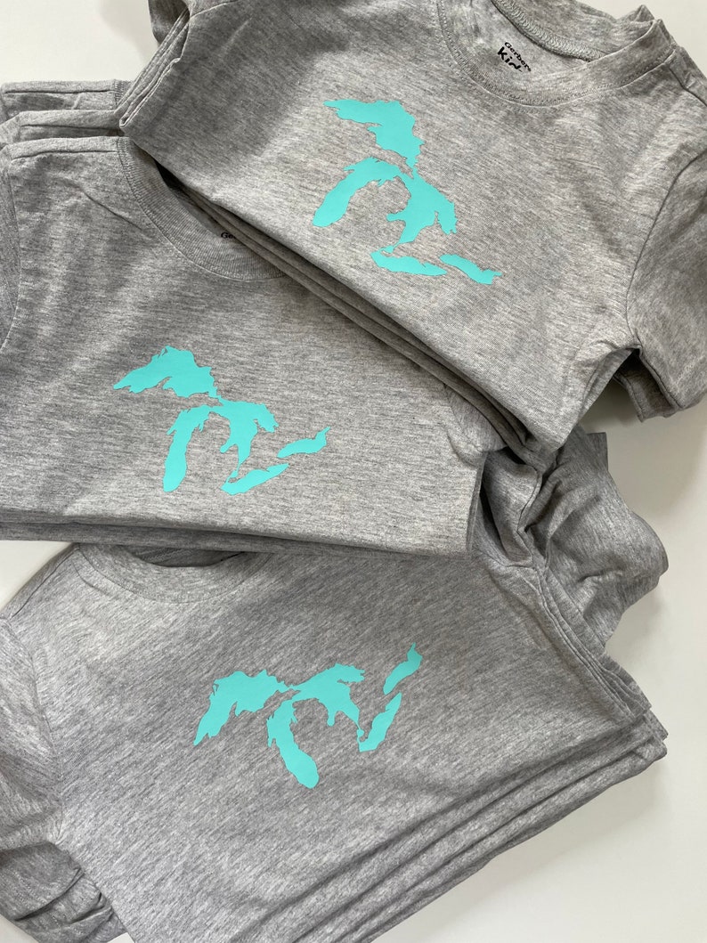 May include: A stack of grey t-shirts with a turquoise blue outline of the Great Lakes on each shirt.