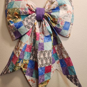 May include: A large, colorful quilted bow with a patchwork design. The bow features squares of fabric in various colors and patterns, including floral prints and bunny motifs. A purple band secures the center of the bow. The bow is a decorative item.