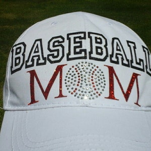 Puede incluir: Gorra de béisbol blanca con las palabras "BASEBALL MOM" en letras negras y rojas. Una pelota de béisbol de diamantes de imitación está centrada entre las palabras.