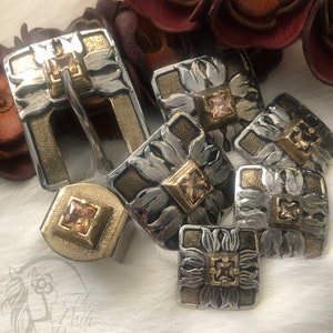 May include: A set of six silver and gold belt buckles with intricate floral designs and clear gemstones. The buckles are arranged on a white furry surface.