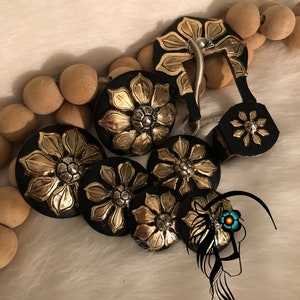 May include: A collection of black and gold floral-designed buttons and a belt buckle. The buttons and buckle feature a gold flower design on a black background. The buckle has a silver metal clasp. The items are arranged on a white, fluffy surface.