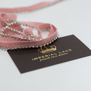 Könnte beinhalten: Rosa Stoffbesatz mit silbernen Perlenrand. Der Besatz ist aufgerollt und eine Visitenkarte mit dem Text "IMPERIAL LACE french lace dealer" ist sichtbar.