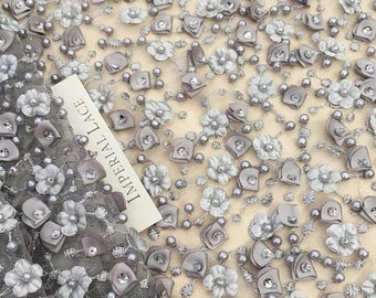 May include: Grey floral lace fabric with 3D flowers and pearl beads. The fabric has a delicate, intricate design and is perfect for adding a touch of elegance to any project.
