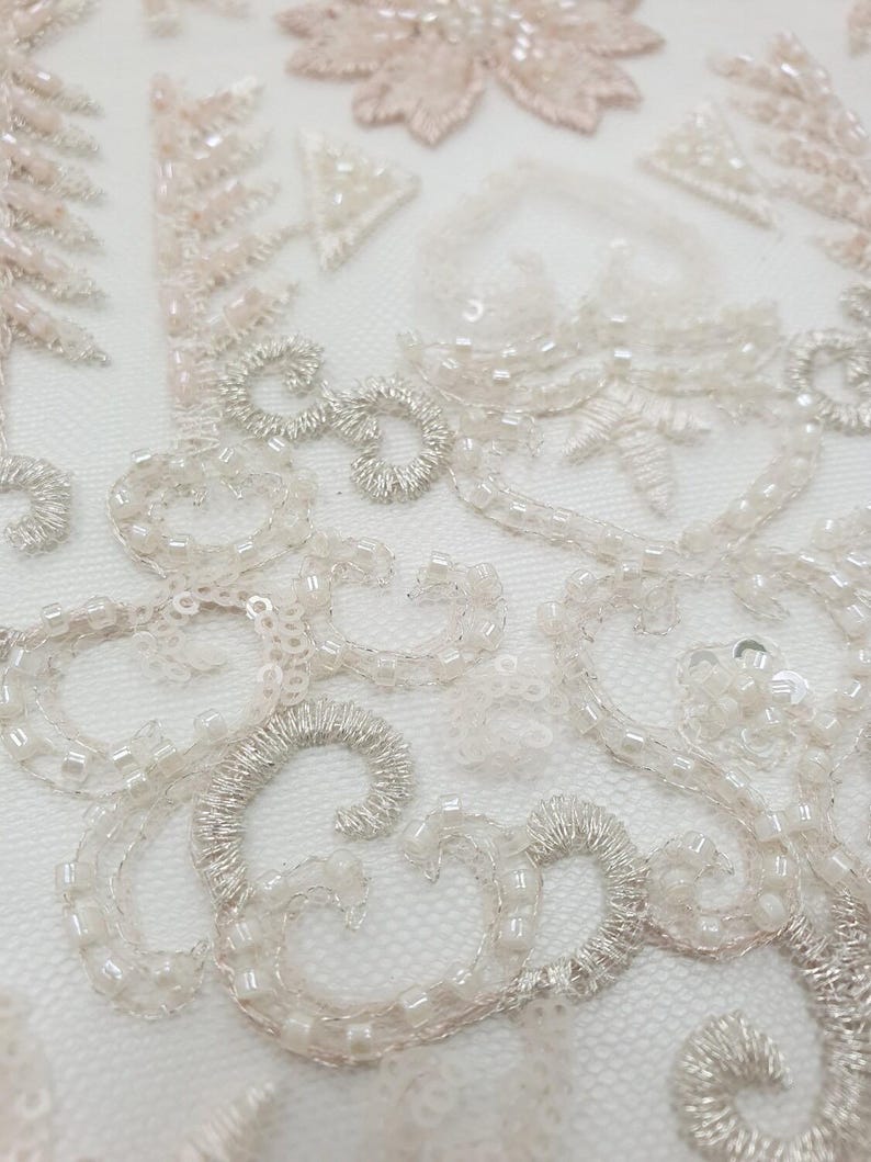 May include: Close-up of delicate white and pink beaded lace fabric with a floral design. The fabric is made of a sheer mesh material and features intricate beading and embroidery.