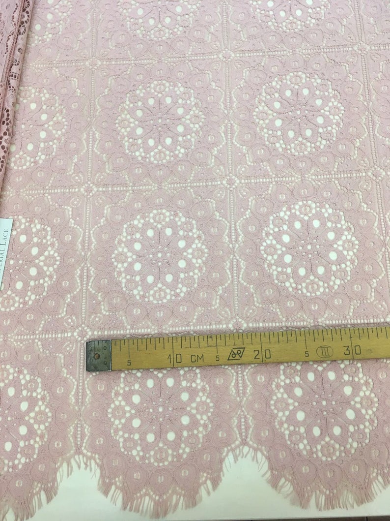May include: Pink lace fabric with a delicate floral pattern. The fabric has a scalloped edge and is measured with a ruler in inches.