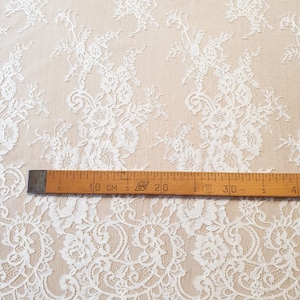 May include: White lace fabric featuring an intricate floral pattern with scalloped edges. A wooden ruler is placed horizontally across the fabric, showing measurements in centimeters, with the 10 cm mark aligned near the left edge of the ruler.
