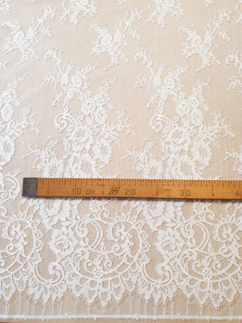 May include: White lace fabric featuring an intricate floral pattern with scalloped edges. A wooden ruler is placed horizontally across the fabric, showing measurements in centimeters, with the 10 cm mark aligned near the left edge of the ruler.