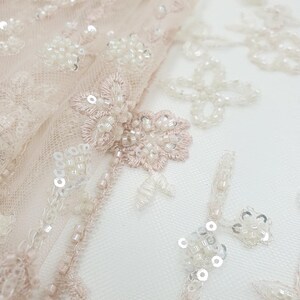 May include: Close-up of delicate blush pink tulle fabric with intricate floral embroidery and white beads and sequins.