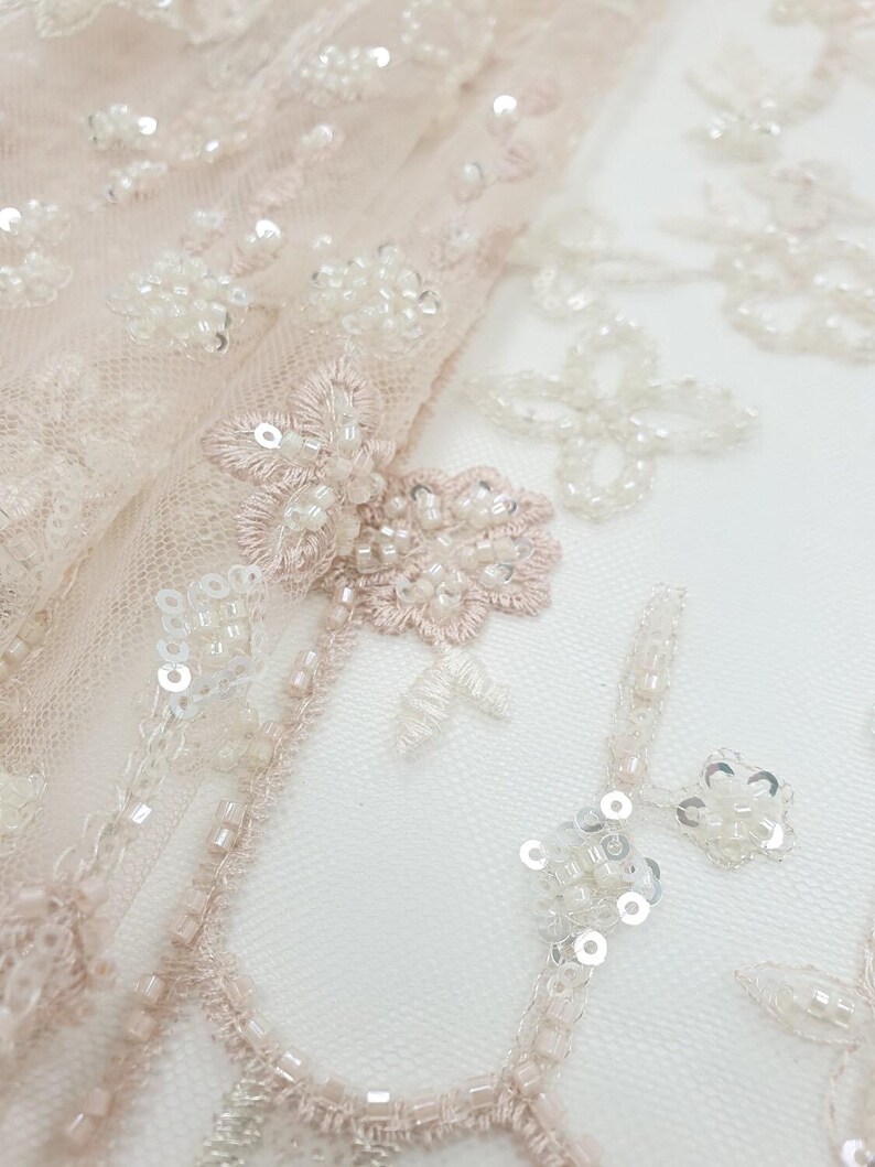 May include: Close-up of delicate blush pink tulle fabric with intricate floral embroidery and white beads and sequins.