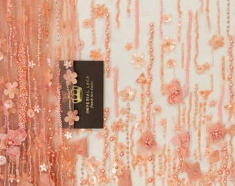May include: Peach-colored beaded lace fabric with floral embellishments. The fabric features a delicate mesh base with rows of beads and sequins, creating a shimmering effect. The floral embellishments are made of soft, layered fabric and are scattered throughout the fabric.