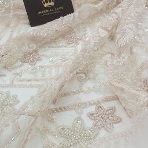 May include: Close-up of delicate, beaded lace fabric with a floral pattern. The fabric is a light pink colour with white beads and sequins. The text "IMPERIAL LACE French lace dealer" is visible on a black card.