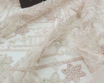 May include: Close-up of delicate, beaded lace fabric with a floral pattern. The fabric is a light pink color with white beads and sequins. The text "IMPERIAL LACE French lace dealer" is visible on a black card.