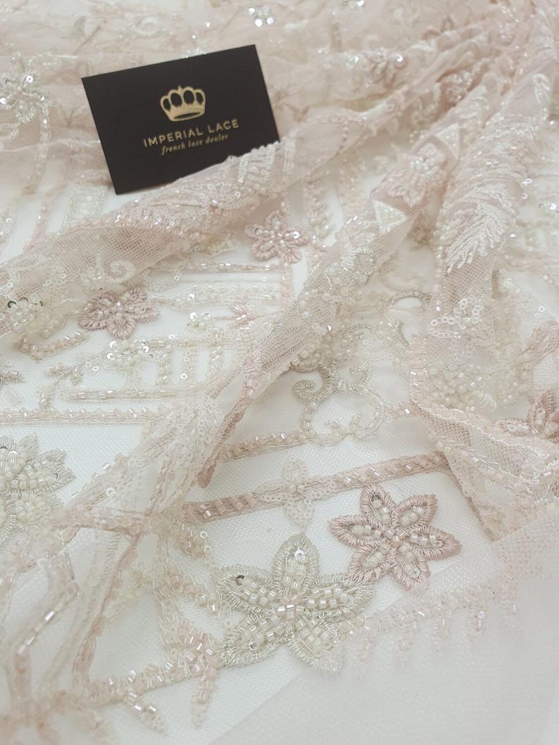 May include: Close-up of delicate, beaded lace fabric with a floral pattern. The fabric is a light pink colour with white beads and sequins. The text "IMPERIAL LACE French lace dealer" is visible on a black card.