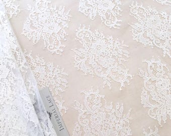 Puede incluir: Primer plano de tela de encaje blanco con patrones florales. El encaje es delicado y detallado, con un fondo blanco roto. Se ve una etiqueta que dice "IMPERIAL LACE". Esta tela es adecuada para vestidos de novia y otras prendas especiales.