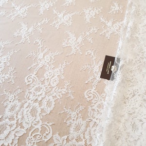 May include: A white lace fabric featuring a delicate floral pattern with intricate embroidery on a sheer mesh base. A small rectangular dark brown tag is attached to the fabric, displaying the text IMPERIAL LACE and a crown icon.