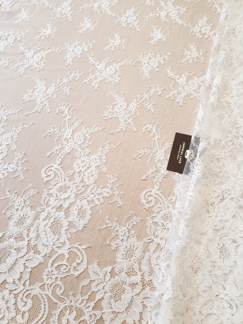 May include: A white lace fabric featuring a delicate floral pattern with intricate embroidery on a sheer mesh base. A small rectangular dark brown tag is attached to the fabric, displaying the text IMPERIAL LACE and a crown icon.