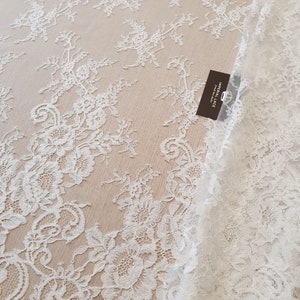 May include: White lace fabric featuring an intricate floral pattern with scalloped edges. A small rectangular black tag is attached to the fabric, displaying the white text IMPERIAL LACE french lace dealer in a vertical orientation.