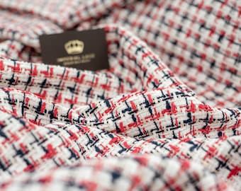 May include: Close-up of a textured fabric with a woven pattern in white, red, and navy blue. The fabric is draped, creating soft folds. A small brown card with a gold crown logo is partially visible.