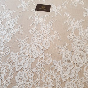 May include: White floral lace fabric featuring intricate rose and leaf patterns on a sheer beige mesh background. A small dark rectangular tag with a gold crown icon and the text IMPERIAL LACE FRENCH LACE DEALER is placed on the fabric.