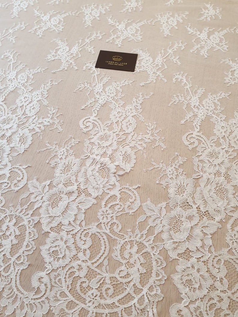 May include: White floral lace fabric featuring intricate rose and leaf patterns on a sheer beige mesh background. A small dark rectangular tag with a gold crown icon and the text IMPERIAL LACE FRENCH LACE DEALER is placed on the fabric.