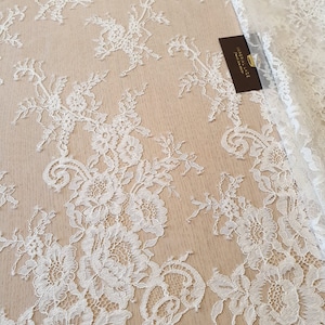 May include: White floral lace fabric featuring an intricate botanical pattern with detailed petals and leaves. A small dark brown rectangular tag with the gold text IMPERIAL LACE is tucked into the folds of the white lace textile.