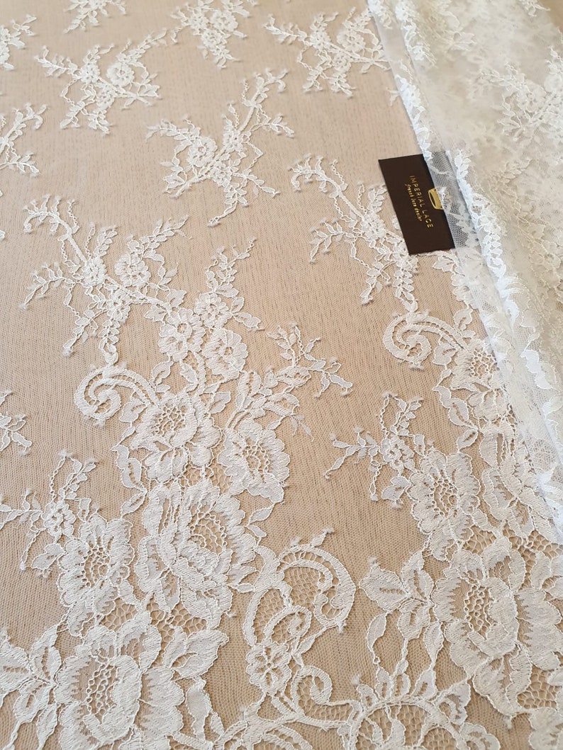 May include: White floral lace fabric featuring an intricate botanical pattern with detailed petals and leaves. A small dark brown rectangular tag with the gold text IMPERIAL LACE is tucked into the folds of the white lace textile.