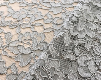 May include: Close-up of grey floral lace fabric. The lace features a detailed pattern of flowers and leaves, with a delicate, open weave. The fabric is draped, revealing the intricate design and texture.