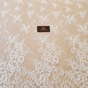 May include: A white lace fabric featuring an intricate floral pattern with scalloped edges and a sheer mesh base. A small rectangular black tag with a gold crown icon and the text IMPERIAL LACE is placed in the center of the fabric.